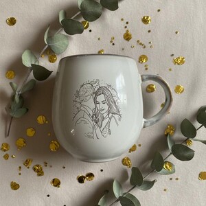 May include: A white ceramic mug featuring a black line art illustration of a couple embracing. The mug has a rounded, bulbous shape with a curved handle on the right side. The design depicts two people in a close, intimate pose.
