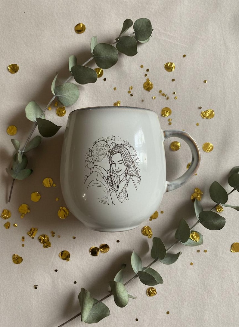 May include: A white ceramic mug featuring a black line art illustration of a couple embracing. The mug has a rounded, bulbous shape with a curved handle on the right side. The design depicts two people in a close, intimate pose.