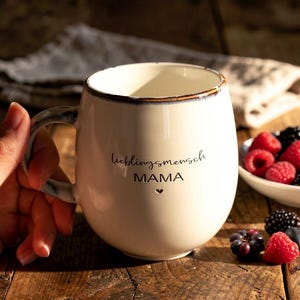 May include: Cream ceramic mug with the words "lieblingsmensch MAMA" and a heart symbol. The mug has a brown rim and handle. Fresh berries are in a white bowl and scattered on a wooden surface.