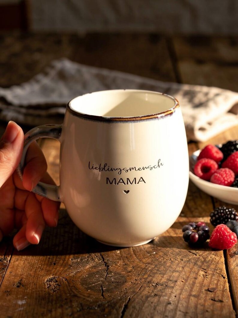 May include: Cream ceramic mug with the words "lieblingsmensch MAMA" and a heart symbol. The mug has a brown rim and handle. Fresh berries are in a white bowl and scattered on a wooden surface.