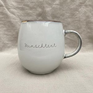 May include: A white ceramic mug with a rounded shape and a gray handle. The rim has a brown accent. The mug has the word "Wunschtext" written on it in a handwritten style. The mug is sitting on a neutral-colored fabric.