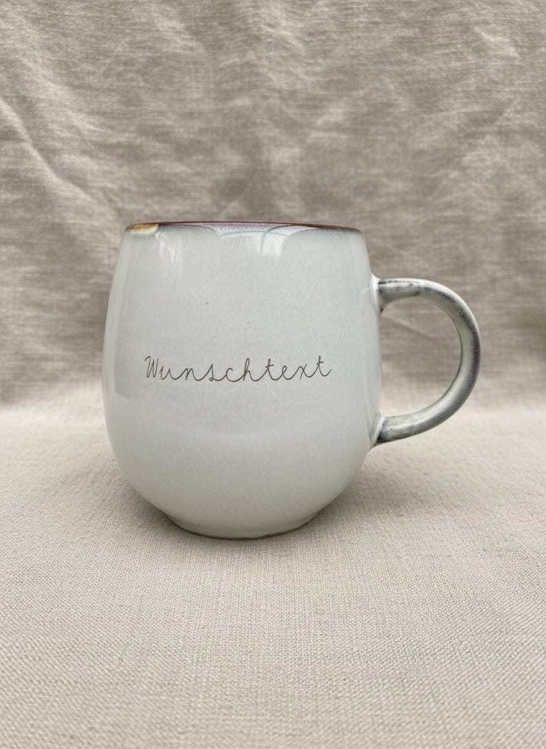 May include: A white ceramic mug with a rounded shape and a gray handle. The rim has a brown accent. The mug has the word "Wunschtext" written on it in a handwritten style. The mug is sitting on a neutral-colored fabric.