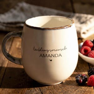 May include: Cream ceramic mug with a rounded shape, dark rim and handle. The mug features the words "Lieblingsmensch AMANDA" and a heart symbol. Berries are in a small white bowl, on a wooden surface.