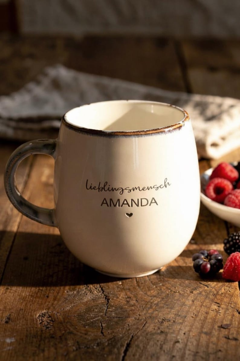 May include: Cream ceramic mug with a rounded shape, dark rim and handle. The mug features the words "Lieblingsmensch AMANDA" and a heart symbol. Berries are in a small white bowl, on a wooden surface.