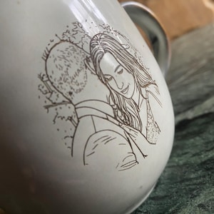 May include: A white ceramic mug featuring a dark brown line art illustration of two people embracing. The drawing depicts one person with short hair and another with long hair, positioned close together in a side profile view on the curved surface.