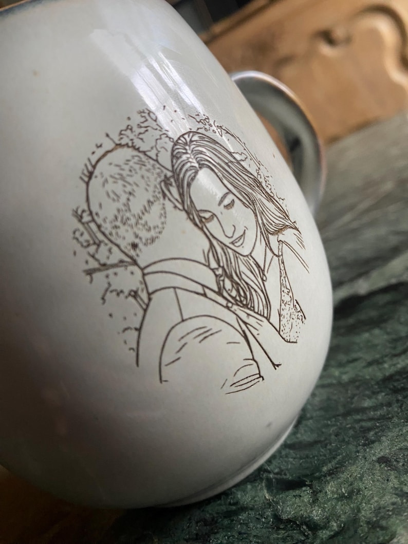 May include: A white ceramic mug featuring a dark brown line art illustration of two people embracing. The drawing depicts one person with short hair and another with long hair, positioned close together in a side profile view on the curved surface.