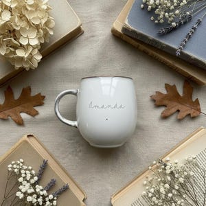 May include: White ceramic mug with the name "Amanda" in script, a small heart, and a gray handle. The mug is surrounded by old books, dried flowers, and fall leaves on a neutral-toned surface.