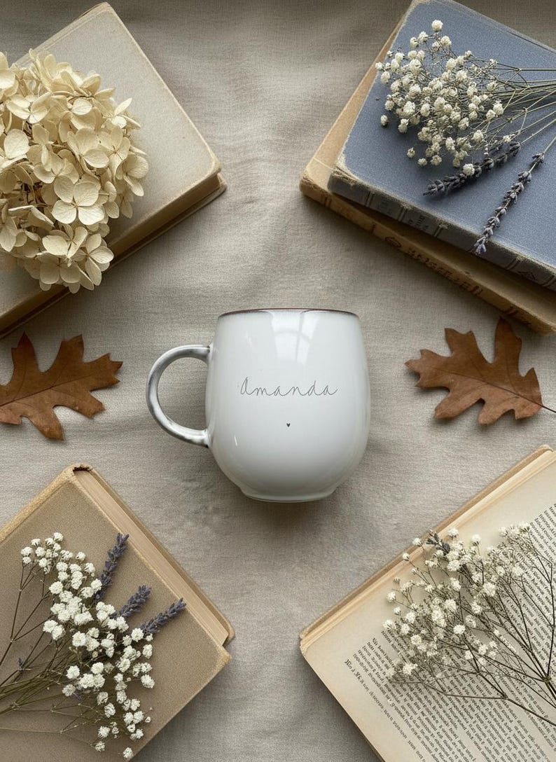 May include: White ceramic mug with the name "Amanda" in script, a small heart, and a gray handle. The mug is surrounded by old books, dried flowers, and fall leaves on a neutral-toned surface.