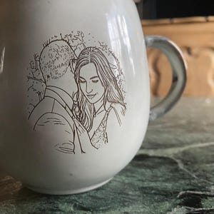 May include: A white ceramic mug featuring a dark brown line art illustration of a couple embracing. The mug has a rounded shape with a smooth finish and a curved handle on the right side. The illustration depicts two people in a close pose.