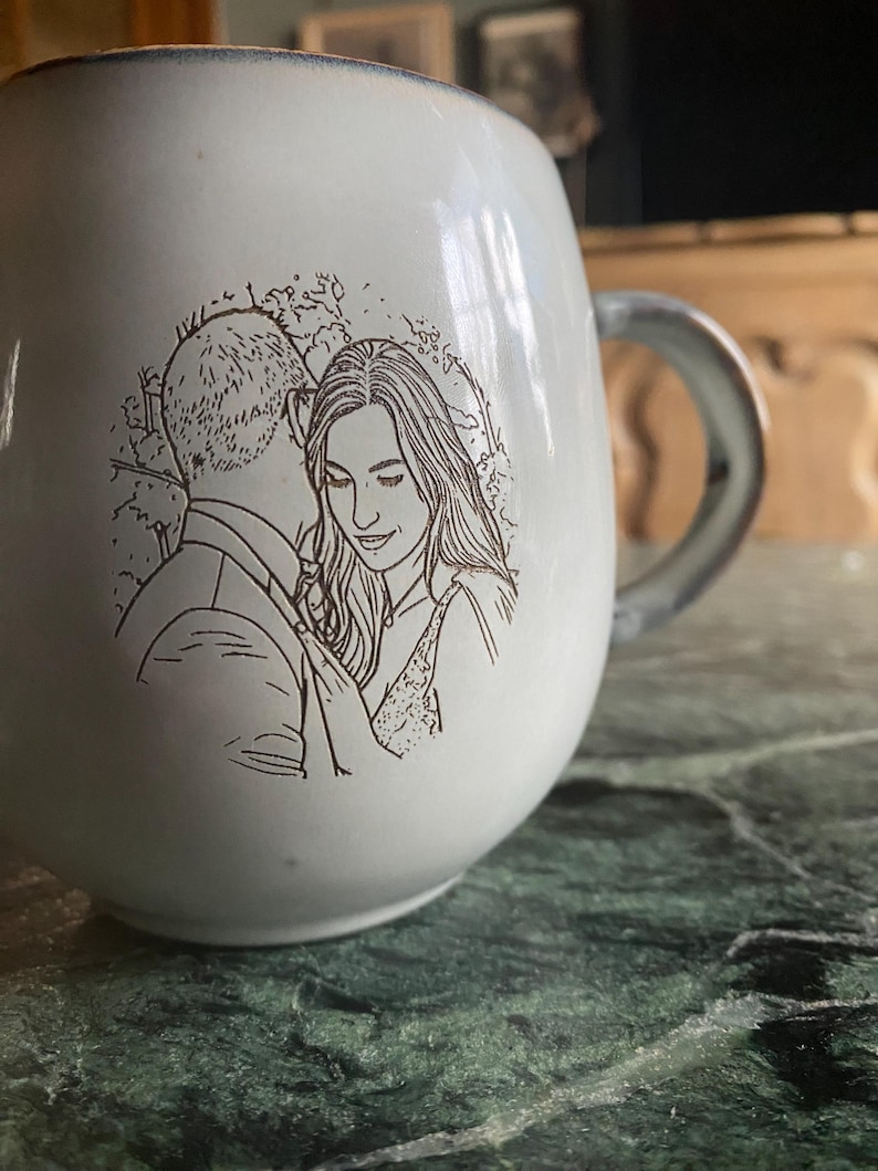 May include: A white ceramic mug featuring a dark brown line art illustration of a couple embracing. The mug has a rounded shape with a smooth finish and a curved handle on the right side. The illustration depicts two people in a close pose.