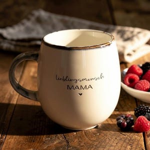May include: Cream ceramic mug with a rounded shape and a dark blue handle. The mug has the words "lieblingsmensch MAMA" and a heart symbol printed on it. Berries are in a small white bowl next to the mug.