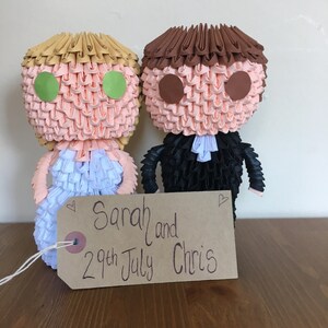 May include: Two origami figures of a bride and groom, with peach-colored faces and green and brown eye details. The bride wears a white and blue dress, and the groom wears a black suit. A tag reads "Sarah and 29th July Chris."