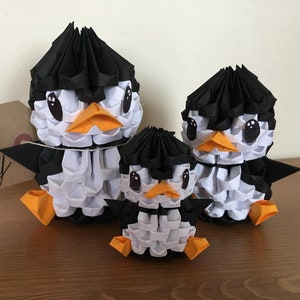 Könnte beinhalten: Drei Origami-Pinguinfiguren in Schwarz, Weiß und Orange. Die Pinguine haben schwarze Köpfe, weiße Körper und orangefarbene Schnäbel und Füße. Die Figuren sind in einer Familiengruppe angeordnet, mit einem großen, mittleren und kleinen Pinguin.