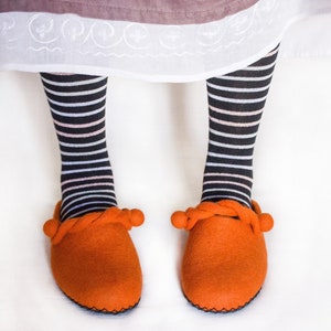 May include: A pair of orange felt slippers with a rope detail and pom poms. The slippers are worn by a person wearing black and white striped socks.