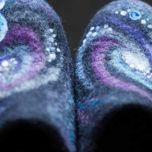 May include: A pair of dark blue wool slippers with a galaxy-themed design. The slippers feature white, blue, and purple swirls and dots, resembling a nebula.
