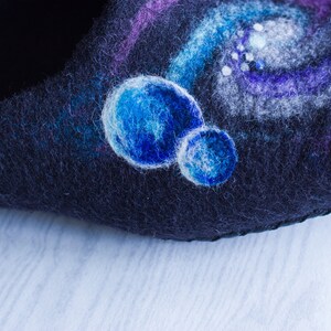 May include: Close-up of a dark blue felt slipper with a hand-painted design of a galaxy with white and blue stars and planets.