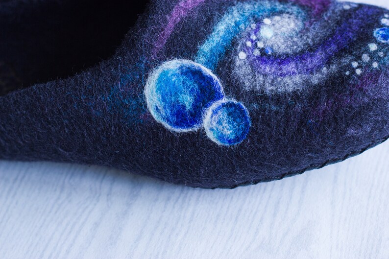 May include: Close-up of a dark blue felt slipper with a hand-painted design of a galaxy with white and blue stars and planets.