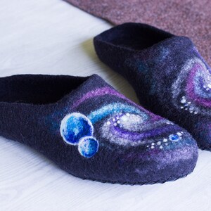 May include: A pair of dark blue wool slippers with a galaxy design. The slippers have a swirling pattern of purple, white, and blue, with small white dots and blue circles.