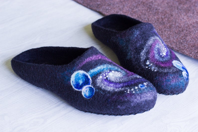 May include: A pair of dark blue wool slippers with a galaxy design. The slippers have a swirling pattern of purple, white, and blue, with small white dots and blue circles.