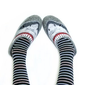 Pu&ograve; includere: Un paio di pantofole in feltro grigio con un design a forma di testa di squalo. Le pantofole sono indossate su piedi con calze a righe bianche e nere.