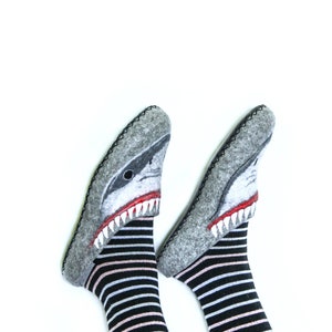 Pu&ograve; includere: Un paio di pantofole in feltro grigio con un design a forma di testa di squalo. Le pantofole sono indossate su piedi con calze a righe bianche e nere.