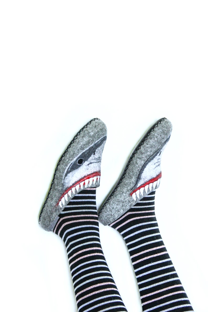 Pu&ograve; includere: Un paio di pantofole in feltro grigio con un design a forma di testa di squalo. Le pantofole sono indossate su piedi con calze a righe bianche e nere.