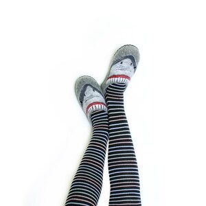 Pu&ograve; includere: Un paio di calze a righe bianche e nere fino al ginocchio con un design a forma di squalo sulle dita. Le calze sono indossate sulle gambe di una persona.