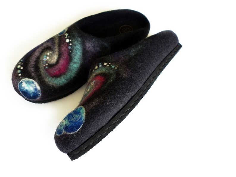 May include: A pair of black felt slippers with a galaxy design. The slippers have a blue, green, and pink swirl pattern with white dots and blue planets.