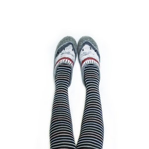 Pu&ograve; includere: Un paio di pantofole grigie a forma di squalo con denti rossi e occhi bianchi. Le pantofole sono indossate con calze a righe bianche e nere fino al ginocchio.