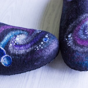 May include: A pair of black felt slippers with a galaxy design. The slippers feature a swirling pattern of purple, blue, and white, with small white dots and larger blue circles.