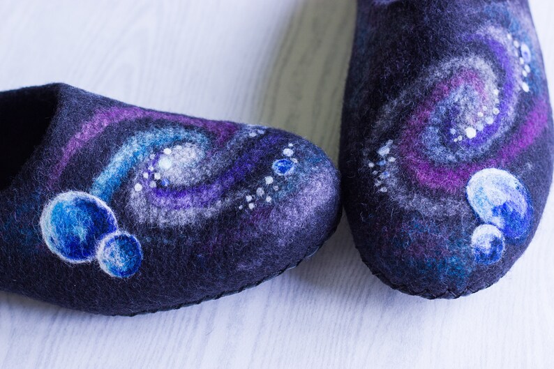 May include: A pair of black felt slippers with a galaxy design. The slippers feature a swirling pattern of purple, blue, and white, with small white dots and larger blue circles.