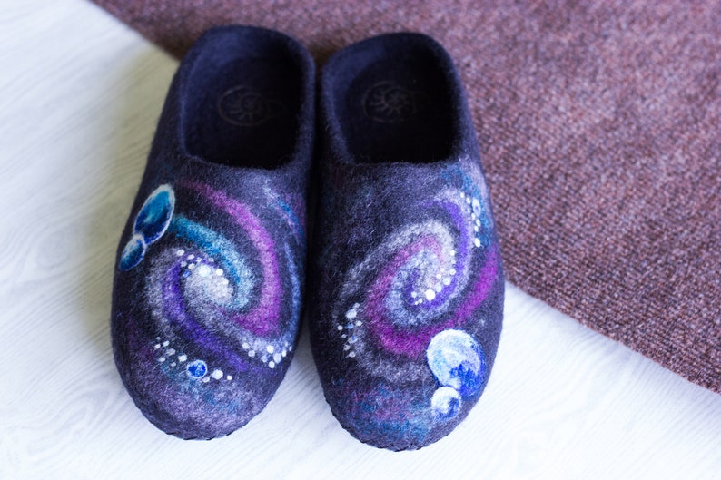 May include: A pair of dark blue wool slippers with a galaxy design. The slippers have a swirling pattern of purple, blue, and white, with small white dots representing stars.