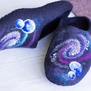 May include: A pair of black felt slippers with a galaxy design. The slippers have a swirling pattern of purple, blue, and white, with small white dots scattered throughout. There are also three blue circles painted on each slipper, representing planets.