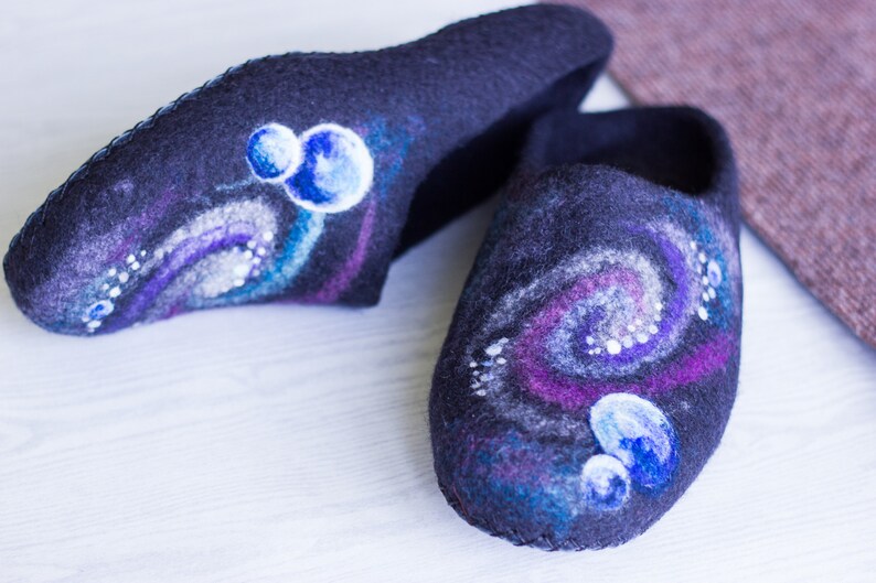 May include: A pair of black felt slippers with a galaxy design. The slippers have a swirling pattern of purple, blue, and white, with small white dots scattered throughout. There are also three blue circles painted on each slipper, representing planets.
