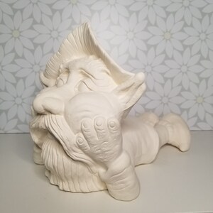 May include: White ceramic gnome figurine with a beard and a hand holding an apple. The gnome is lying on its side with its head resting on the apple.