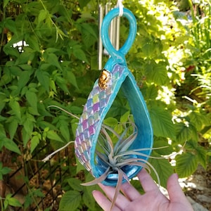 Könnte beinhalten: Ein türkisfarbener Keramik-Hängepflanztopf mit geometrischem Muster und goldener Akzent. Der Topf ist tropfenförmig und hat oben eine Schlaufe zum Aufhängen. Eine kleine Luftpflanze wächst im Topf.