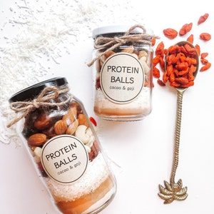 May include: Two glass jars filled with ingredients for protein balls. The jars are labeled "PROTEIN BALLS cacao & goji". One jar has almonds, cashews, dried cranberries, and shredded coconut. The other jar has almonds, cashews, dried goji berries, and shredded coconut. A gold spoon with a palm tree design is in the background.