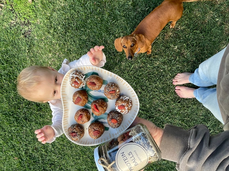 May include: A heart-shaped plate with nine protein balls topped with goji berries, coconut flakes, and chia seeds. A glass jar with a label that reads "Protein Balls" is in the foreground. A baby and a dachshund dog are in the background.