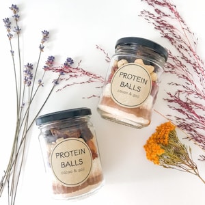 May include: Two glass jars filled with ingredients for protein balls. The jars have brown paper labels with the text "PROTEIN BALLS cacao & goji".
