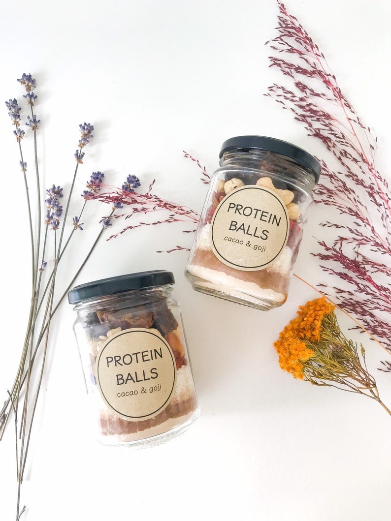 May include: Two glass jars filled with ingredients for protein balls. The jars have brown paper labels with the text "PROTEIN BALLS cacao & goji".
