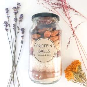 May include: A glass jar filled with ingredients for protein balls. The jar has a brown label that says "PROTEIN BALLS cacao & goji".
