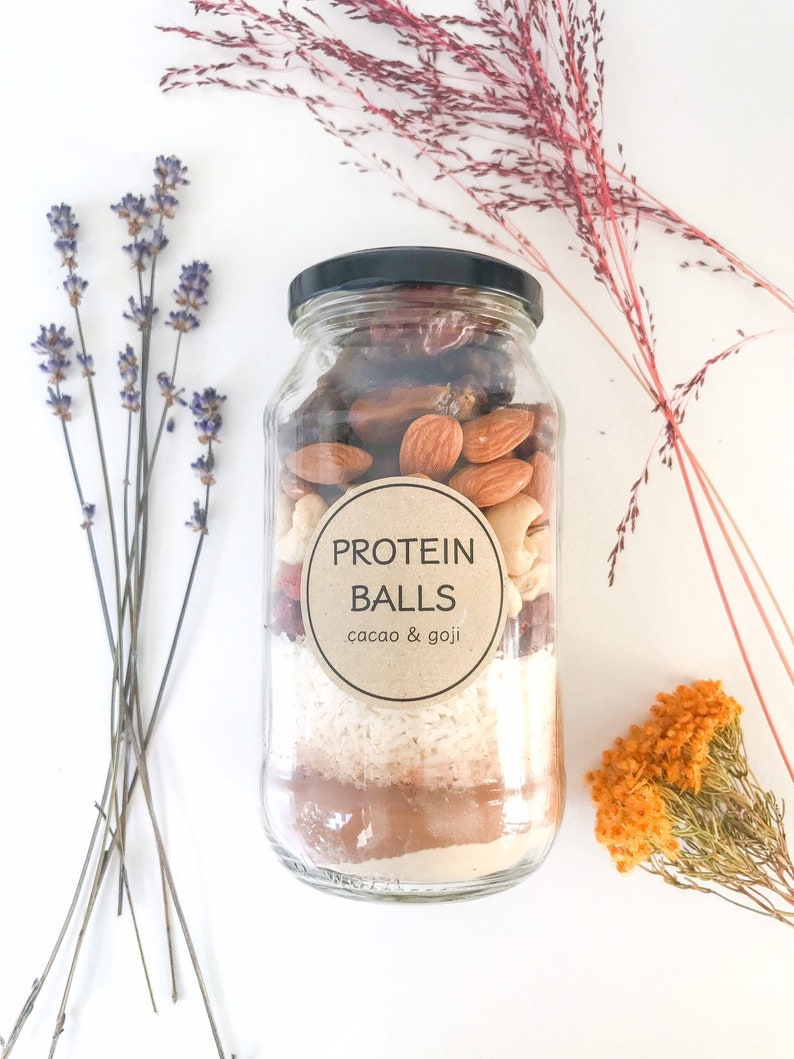 May include: A glass jar filled with ingredients for protein balls. The jar has a brown label that says "PROTEIN BALLS cacao & goji".