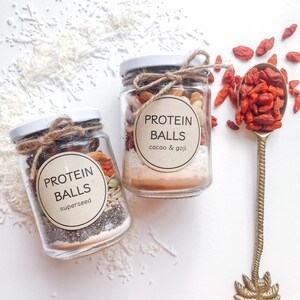 May include: Two glass jars filled with various ingredients, including nuts, seeds, and dried fruit. The jars are labeled "Protein Balls" with different flavor variations: "cacao & goji" and "superseed".