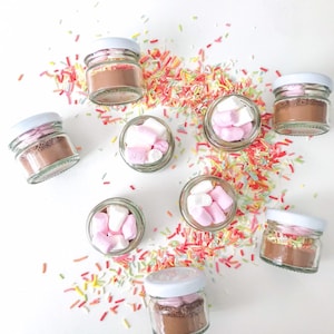 May include: A collection of small glass jars filled with hot chocolate mix and marshmallows. The jars are scattered on a white surface with colourful sprinkles.