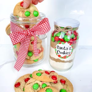 May include: Two glass jars filled with ingredients for Santa's Cookies. One jar is filled with cookies and tied with a red and white checked ribbon. The other jar has a label that says "Santa's Cookies" and is filled with ingredients and candies. Several cookies with red and green candies are in front.