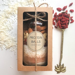 May include: A brown cardboard box with a clear window showing a glass jar filled with protein balls. The jar is tied with twine and has a label that reads "PROTEIN BALLS cacao & goji". The box is sitting on a white surface with shredded coconut and dried goji berries.