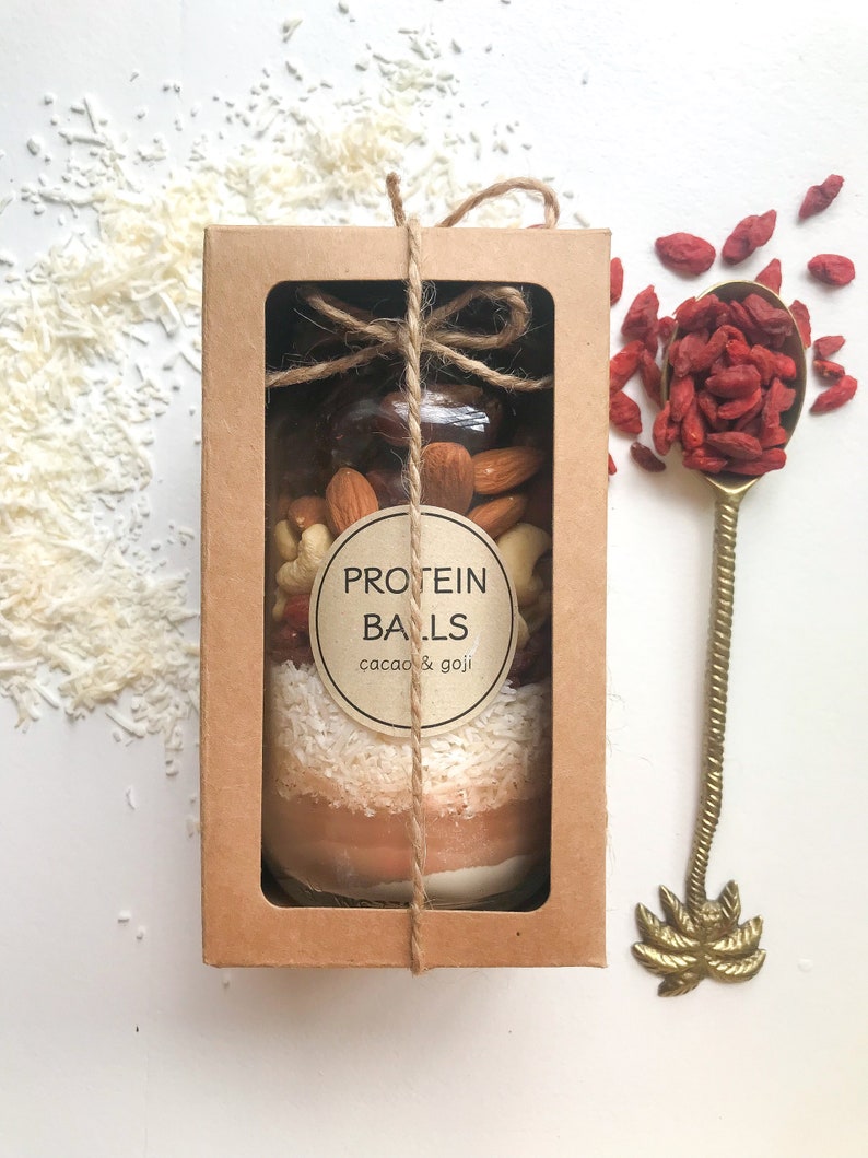 May include: A brown cardboard box with a clear window showing a glass jar filled with protein balls. The jar is tied with twine and has a label that reads "PROTEIN BALLS cacao & goji". The box is sitting on a white surface with shredded coconut and dried goji berries.