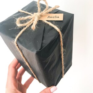 May include: A black gift box wrapped in brown paper and tied with twine. A brown paper tag with the name "Amelia" is attached to the twine.
