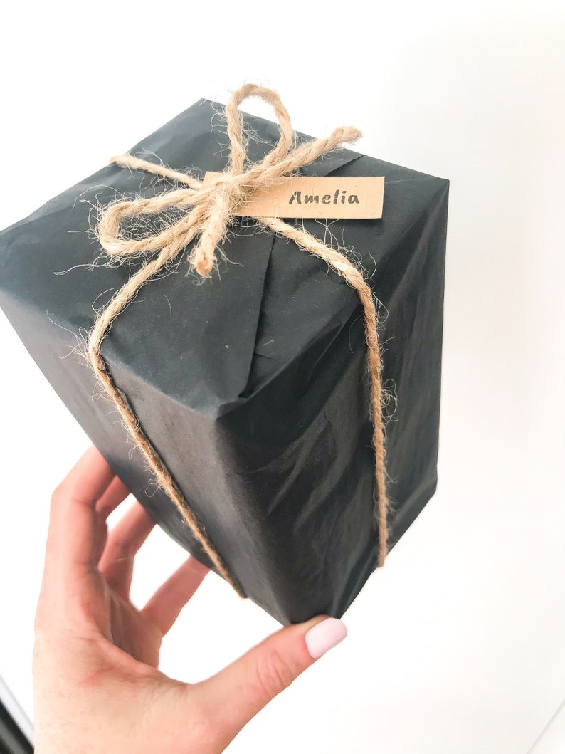 May include: A black gift box wrapped in brown paper and tied with twine. A brown paper tag with the name "Amelia" is attached to the twine.