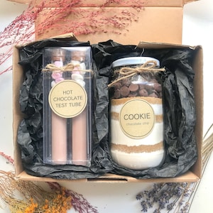 May include: A brown cardboard box with black tissue paper inside. The box contains two glass test tubes filled with a pink and white substance labelled "Hot Chocolate Test Tube" and a glass jar filled with cookie ingredients labelled "Cookie Chocolate Chip".